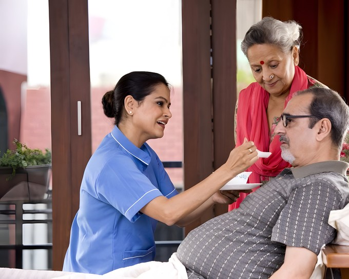 Nurse helping an elderly man with his meal while his wife looks on at home